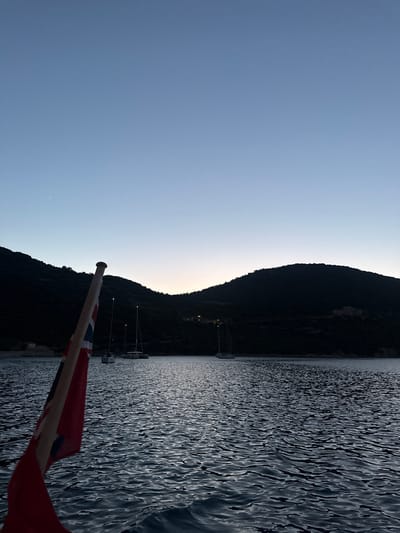 A British flag, unfurled and partially visible in the foreground, flies from a flagpole on a boat. The flag's red, white, and blue pattern is distinct. The boat is on the water, which exhibits a rippling texture, suggesting movement. In the mid-ground, several sailboats are anchored or moored. Beyond the sailboats, a dark, hilly coastline rises, partially obscured by the fading light of dusk. A few lights are visible on the hillside, suggesting a settlement. The sky is a gradient of pale blue, transitioning to a warmer hue near the horizon, indicating twilight. A single bright star or planet is visible in the upper left portion of the sky. The scene is peaceful and serene, likely captured in the bay of Vathy, Greece, a common location for sailing and anchoring due to its sheltered waters and picturesque surroundings.