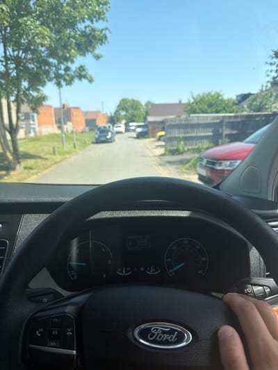 A first-person perspective from the driver's seat of a Ford vehicle, likely a van, is depicted. The dashboard and steering wheel are prominently visible. The speedometer reads approximately 25 mph. The vehicle appears to be driving down a street lined with houses with red roofs. The location is likely in a residential area in Swindon, United Kingdom. A red vehicle is visible on the side of the road. A hand is visible on the steering wheel.
