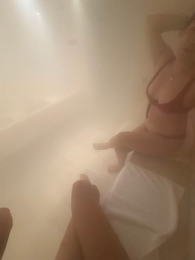 A woman is partially submerged in a bathtub, wearing a dark-colored bikini top and draped with a white towel. Her hand is reaching out, seemingly touching the water. The scene has a hazy, foggy appearance. There is no visual evidence to confirm the location as Pueblo Nuevo de Jasso, Mexico.