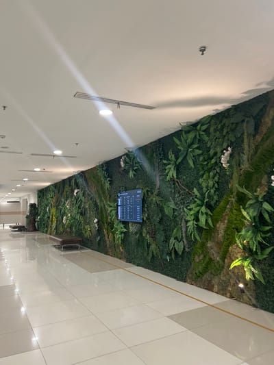 The image shows a long, curved hallway with a living green wall on the right side. The wall is adorned with various plants, including ferns and white flowers, and appears to be a decorative feature. Mounted on the green wall is a digital display screen showing information, likely flight or boarding details. Along the left side of the hallway, there is a polished tiled floor that reflects the light from the ceiling. Several brown benches are placed in the hallway, two of which are visible in the foreground and mid-ground. The ceiling is white and features recessed lighting and air vents. The overall impression is of a modern, well-maintained indoor space with a touch of nature integrated into the design. The scene is likely located in an airport or a similar public transportation hub within the city of Baji Mangngai, Indonesia, given the presence of the digital display and the seating arrangements typical of such locations.