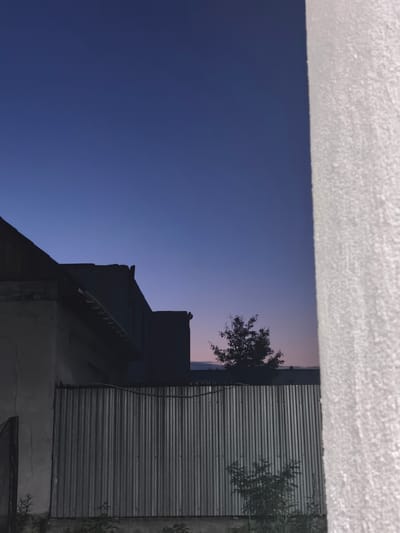 The image depicts a twilight scene in what appears to be a residential or semi-rural area. A large, rough-textured white wall dominates the right side of the frame. To the left, silhouetted against a deep blue sky transitioning to pale pink at the horizon, are the rooftops of buildings and a corrugated metal fence. A lone tree is visible in the middle ground, its branches stark against the fading light. The overall impression is one of quiet dusk, with a focus on architectural and natural elements. The location is identified as Белоярская Застава, Russia.