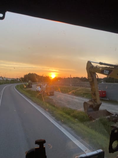 The image shows a construction site at sunset in Podłęże, Poland. A yellow CAT excavator is the prominent object in the foreground, with its arm extended. Beyond the excavator, a road curves to the right, bordered by a grassy verge and trees. The sky is filled with the warm hues of sunset, casting a golden light over the scene. A few small vehicles are visible in the distance. The overall impression is one of industrial activity at the end of the day. The time of day is clearly sunset, indicated by the low angle of the sun and the warm colors in the sky. The weather appears to be clear. The location is specified as Podłęże, Poland. No people are visible in the image. The text "CAT" is visible on the excavator.