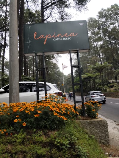 In Cikole, Indonesia, a prominent green sign for "lapinea CAFÉ & RESTO" stands beside a road surrounded by tall trees and lush vegetation. The sign, featuring orange cursive text, is supported by a dark metal frame. In the foreground, a vibrant bed of orange marigolds bursts with color. On the right side of the road, a silver car is parked, and on the left, a white car is partially visible. The road itself shows light traffic, with a few other vehicles and a person walking in the distance. The overall atmosphere suggests a scenic, possibly mountainous, location that caters to travelers seeking refreshment.