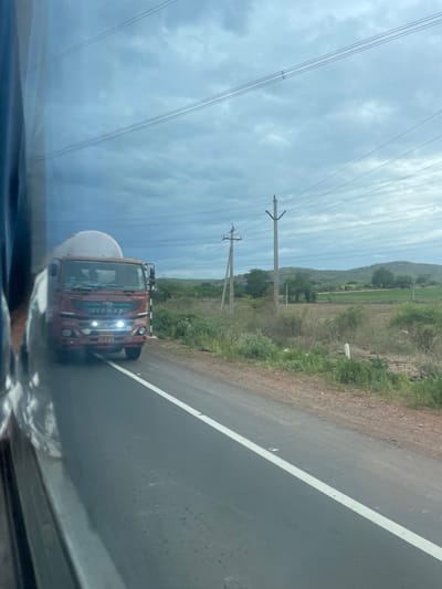 In Sugali Tanda, India, a red Eicher truck, possibly transporting liquid in a large white cylindrical tank, is traveling on a paved road under a cloudy sky. The truck has its headlights on. The road has a white line marking its edge. In the background, there are fields, trees, hills, and electrical utility poles with overhead wires. The image is taken from inside a vehicle, as indicated by the reflection of its interior on the left side of the frame.