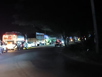The image depicts a night scene on a road in Keudetanjong, Indonesia. A line of vehicles, including several tanker trucks and cars, are parked along the left side of the road. The tankers have hazard identification markings on their rear. Some of the vehicles have their headlights on. The right side of the road is lined with trees and vegetation, casting shadows and obscuring detail. A few other vehicles are visible further down the road. The overall lighting is poor, with light sources limited to vehicle headlights and some street lights in the distance.