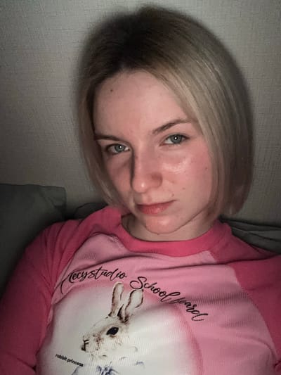 A medium-shot portrait depicts a young individual, likely female, lying down indoors. The person has short, light blonde hair with visible darker roots and light blue-green eyes, looking directly at the camera with a neutral to slightly contemplative expression. They are wearing a two-tone long-sleeved top, primarily pink, with a lighter pink or white central panel.
The top features a graphic of a white rabbit. Several lines of text are visible on the garment: "Mecy studio School yard" is written in a cursive, dark script above and to the left of the rabbit graphic, and "rabbit princess" is printed in smaller, dark text below the rabbit.
The background consists of a textured grey wall and what appears to be grey bedding or upholstery, suggesting a casual indoor environment such as a bedroom or living room. The lighting is dim and low-key, indicating an evening or night time setting. The overall mood of the image is relaxed and informal. The scene is located in Ligolambi, Russia.
