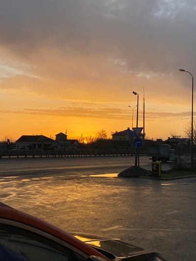 A sunset casts an orange glow across a street and sky. The scene includes streetlights, road signs, and buildings silhouetted against the vibrant sky. The road is wet, reflecting the warm light. A portion of a car is visible in the foreground, adding to the perspective of the street. The sky blends from gray clouds to bright orange near the horizon.