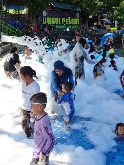 A group of people are playing in a pool filled with white foam. The pool is located at the water park "Umbul Pelem" in Tulung, Indonesia (latitude: -7.592, longitude: 110.65). The sign "Umbul Pelem - Tulung - Klaten" is visible in the background. There are 10 people in the image. The foam is thick and covers most of the pool. A blue tiled surface can be seen underneath the foam. Several people are wading in the foam, including a young boy wearing blue swim trunks and a girl wearing a pink shirt and a blue swimsuit. An adult woman is wearing a blue hijab and a grey jacket, and appears to be interacting with the young boy. A child is wearing goggles and a purple shirt. The foam is covering the bottom of the pool, and there is a small amount of water visible under the foam.