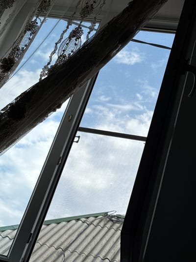 The image shows a view looking up and out of a window with a screen and curtains. The sky is visible with some clouds. A portion of a tiled roof is visible below the window. The scene is not in a city; it is a view from what appears to be a building with a roof. There are no people or discernible actions taking place. The location of Tsiyab-Tsoloda, Russia is irrelevant to the visual content of the image.