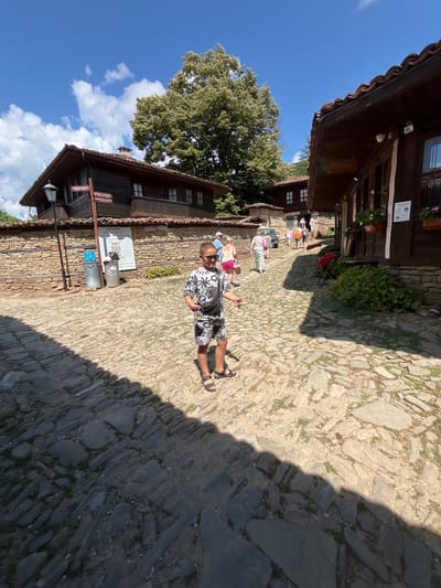 In Zheravna, Bulgaria, a young boy with short, dark hair and sunglasses stands on a cobblestone street, facing the camera. He is wearing a black and white patterned t-shirt and shorts, with black sandals. A black fanny pack is worn across his chest. Behind him, the street is lined with traditional Bulgarian Revival-era houses constructed with stone foundations and dark wooden upper floors, featuring red tile roofs. Several people, seemingly tourists, are visible in the background walking along the street and entering buildings. The sky is blue with scattered white clouds, and a large, leafy tree provides shade. The scene is illuminated by bright sunlight, casting shadows across the cobblestones.