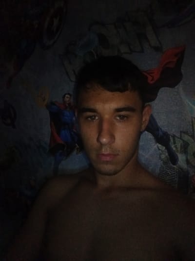 The image depicts a young man, shirtless, with dark hair and brown eyes, looking directly at the camera. He has a faint mustache and light stubble. His skin tone appears to be olive. He is positioned in the foreground, with his upper body taking up most of the frame.
The background is a dark, possibly bedroom wall, adorned with superhero-themed imagery. Elements visible include what appears to be Captain America's shield on the left side, and a depiction of Doctor Strange in his blue and red costume on the right. There are also other superhero-related graphics, partially obscured by the dim lighting and the main subject. The overall lighting is low, casting shadows and contributing to a somewhat moody atmosphere. The setting is not identifiable as a specific city location within Kyzyltog (Krasnogorsk), Uzbekistan, as there are no discernible urban landmarks or environmental cues.