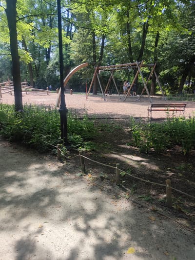 This media file captures a sun-dappled playground scene in a park, likely during a warm, clear day. Tall trees with lush green foliage form a canopy overhead, casting intricate shadows on the sandy ground. In the mid-ground, a wooden swing set stands prominently, with two young girls in casual summer attire appearing to be playing on it. A red rope hangs from one of the swings. To the left, a wooden climbing structure is visible, and further back, a person in a blue shirt stands near some trees. A brown wooden park bench is situated to the right of the swing set, offering a place to rest. A black lamppost is positioned in the foreground, between a patch of dense green bushes and the playground. A simple rope fence with wooden posts delineates a path in the foreground, adding to the park's atmosphere. The overall mood is peaceful and idyllic, suggesting a typical afternoon outing in a public park.