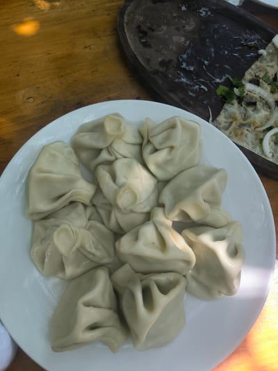 The image shows a white plate filled with khinkali, a type of Georgian dumpling, on a wooden table. Another wooden dish containing the remains of a meal is visible in the background. The location is Karatakla, Georgia.