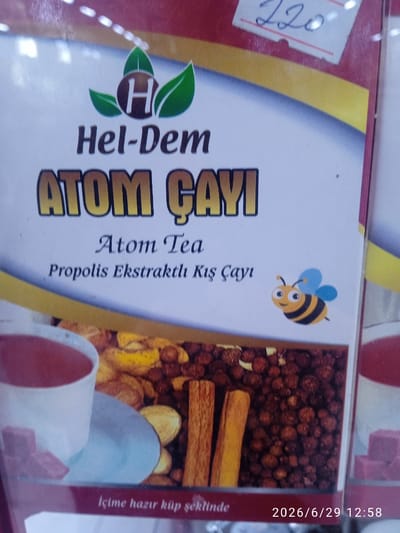 A close-up, slightly blurry photograph shows a product box for "Atom Çayi" (Atom Tea). The box features a logo with the letter "H" inside a circle, flanked by green leaves. Below the logo, the text "Hel-Dem" is visible. The prominent text "ATOM ÇAYI" is displayed in large, stylized letters. Further down, smaller text reads "Atom Tea" and "Propolis Ekstraktlı Kış Çayı," indicating it's a propolis-infused winter tea. A cartoon drawing of a bee is on the right side of the box. The lower portion of the box displays an image of a white teacup with red tea and some pink cubes on a saucer, alongside dried fruits, spices like cinnamon sticks, and peppercorns. A strip along the bottom edge of the box has text that reads "İçime hazır küp şeklinde" and a date stamp "2026/6/29 12:58". The background of the box has a yellow and red gradient. The image appears to be taken in a retail setting in Ak-Suu, Kyrgyzstan, as indicated by the timestamp and context from the user's prompt.