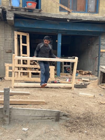 A man is standing in a cluttered outdoor workshop in Dras, India, holding a large wooden frame. He is wearing a dark grey long-sleeved shirt and blue jeans, with orange-soled sandals. The frame appears to be part of a bed or a similar structure. Wood scraps and tools, including a red and black power drill, are scattered on the ground around him. In the background, a workshop structure is visible with more wooden materials and machinery. The scene suggests ongoing construction or woodworking activity in a somewhat unorganized environment.
