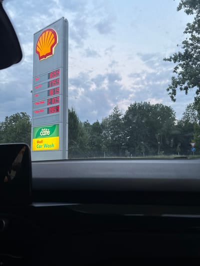 An exterior, eye-level, medium shot captures a Shell gas station sign in Groß-Gerau, Germany, under a cloudy sky. The sign, a tall, vertical structure, prominently displays the Shell logo at the top. Below the logo, a digital display shows fuel prices. From top to bottom, the prices are: Diesel 169.9, Super E10 175.9, Super 181.9, V-Power Racing 210.9, and V-Power Diesel 195.9. Beneath the fuel prices, a green and yellow sign reads "Shell café" and "Shell Car Wash". The foreground is dominated by the interior of a car, with a dark dashboard visible. Trees and foliage form the backdrop to the gas station.