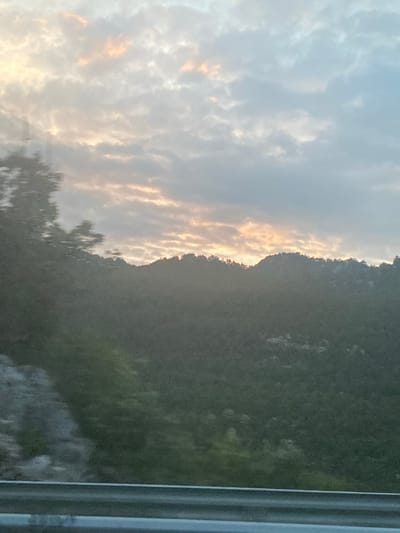 This is a vertical, daytime, outdoor, full shot of a mountain range with a cloudy sky. The sun is setting behind the mountain, casting a warm glow on the clouds. The mountain is covered in lush green trees, and the sky is a mix of blues and whites with streaks of orange and yellow from the setting sun. The overall mood of the image is peaceful and serene, with a sense of awe at the natural beauty of the scene. The image appears to be taken from a moving vehicle, as evidenced by the blurred lines on the right side of the frame, which resemble a window frame.