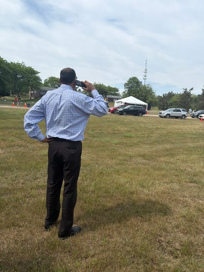 A man in a blue and white checkered button-down shirt and black pants stands in a dry, grassy field, talking on a cell phone held to his right ear. His left hand is on his hip. In the background, there are several vehicles, including a black SUV and a silver SUV, parked in a line. A white tent is set up near the vehicles. Trees and a cell tower are visible in the distance. The sky is partly cloudy. The location is identified as the Town of Southampton, United States of America.