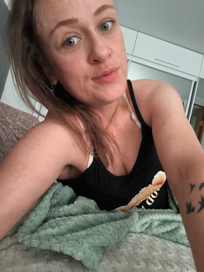 A woman with blonde hair and blue eyes is lying in bed, looking at the camera. She is wearing a black tank top with a white bra underneath. A green blanket is draped over her. In the background, there is a closet with sliding doors. The scene appears to be a bedroom. This image was taken in Kohtla-Järve linn, Estonia.