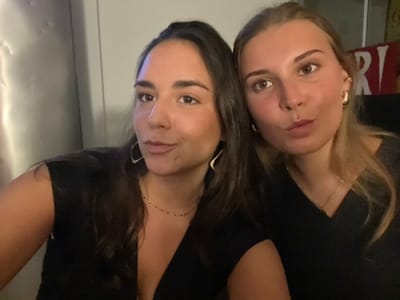 The image displays two adult females in a close-up, chest-up shot within an indoor, dimly lit environment. The female on the left has long, dark brown hair, an olive complexion, and is wearing a black V-neck top, gold-toned geometric hoop earrings, and a delicate dark beaded necklace. She is looking slightly to her right with a subtle smile. The female on the right has long, light brown hair, a fair complexion, and is wearing a black V-neck top, gold-toned hoop earrings, and a delicate gold-toned chain necklace. She is looking directly at the camera with puckered lips. Both subjects are positioned closely together, suggesting a close-range photograph or selfie. The background is dark and indistinct. To the left, a light gray, possibly metallic, textured vertical surface is visible. To the right, a blurry red object with partial white lettering, "RI," is discernible, indicative of an establishment sign. The specific location within the city of Bath, United Kingdom, cannot be determined from the visual evidence present in the image.
