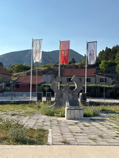 The image shows a monument in the city of Kopito, Montenegro. The monument features a large, abstract stone sculpture with two large curved elements meeting at the top, resting on a rectangular stone base. Three flags are flown on flagpoles to the right of the sculpture. The leftmost flag is white with an emblem in the center. The middle flag is red with a double-headed eagle emblem, which is the national flag of Montenegro. The rightmost flag is white with a blue border and an emblem of a red shield with a white crest. In the background, there are several stone buildings with red tile roofs, surrounded by vegetation and a rocky hillside with trees. The foreground shows a paved courtyard with square stone tiles, some of which are cracked and overgrown with grass. The sky is clear and blue, indicating a daytime scene. There are no people visible in the image.