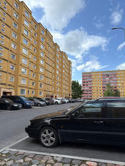 An upward view showcases a row of parked cars beside a street. The foreground is dominated by a black car. Behind the cars are two apartment buildings. The taller building has a stepped, pyramid-like design, and its facade is a bright yellow color with many windows. The second building is a reddish color with yellow accents, and has similar windows. The sky is blue with some white clouds, and a lamppost is also in view.