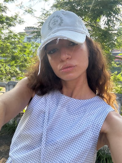 A woman wearing a white baseball cap and a white plaid shirt is in Lao Cai Ward, Vietnam. She is positioned outdoors in a location with trees and a building visible in the background. The woman's face is slightly tilted towards the camera, and her expression is neutral.