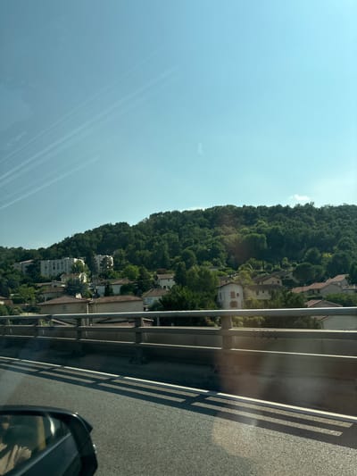 The image captures a view from a vehicle traveling on a road, likely a highway or a major thoroughfare, as indicated by the presence of a guardrail and lane markings. The perspective is from the driver's side, with a portion of the side mirror visible in the lower left corner. The road surface is asphalt, with distinct white lines delineating the lanes.
In the background, a hillside covered in dense green trees rises prominently. Clustered on the slope of this hill are numerous residential buildings, characterized by terracotta tiled roofs and light-colored facades. Some of these buildings appear to be modern apartment blocks, while others are smaller individual houses. The architecture suggests a suburban or semi-rural setting within a temperate climate.
The sky is a clear, bright blue, with some faint, wispy clouds or contrails streaking across the upper portion of the frame, indicating a sunny day. The lighting suggests it is midday or late afternoon, with the sun casting shadows from the guardrail onto the road.
The location is identified as Saint-Romain-en-Gal, France, a commune in the Rhône department. The scene depicts a typical view of a town or city situated against a natural, hilly backdrop, likely captured during transit through the area. There are no people clearly visible in the main part of the image. The overall impression is one of peaceful, sunlit suburban landscape viewed from a moving vehicle.
