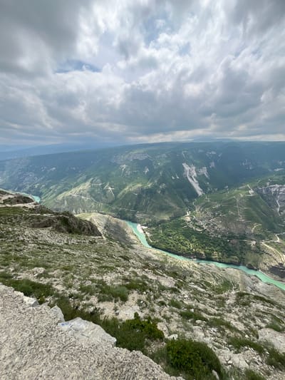 A high-angle, landscape-oriented photograph captures a panoramic view of a rugged landscape near Chirkey, Russia. A wide river, turquoise in color, snakes through a deep canyon. The canyon walls are steep, covered in vegetation ranging from dense forest to sparse grass and rocky terrain. Winding paths and possible roads are visible within the canyon, suggesting human activity or access routes.
The foreground features a rocky cliff edge, with green vegetation growing between the rocks. The sky is overcast with a mix of dark and light clouds. The overall impression is one of dramatic natural beauty and a sense of height and depth.