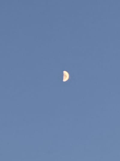 A crescent moon is visible in the sky above Gurkovo, Bulgaria. The moon is illuminated on its right side and appears to be a bright yellow-white. The sky is a clear, pale blue. There are no other celestial bodies or objects visible in the frame.