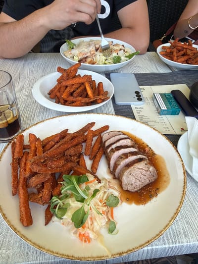 This image depicts a close-up view of a meal served at a restaurant. The primary focus is a white plate with a brown rim, containing a sliced pork tenderloin in a dark sauce, a generous portion of sweet potato fries, and a small salad of shredded cabbage and carrots garnished with lettuce. To the left of this main plate is a glass of dark beverage with lemon slices, and another white bowl filled with more sweet potato fries. In the background, a partially visible person is using a fork to eat from a salad in a white bowl. Another person's arm and shoulder are visible, wearing a black t-shirt. A light gray smartphone, a pack of Terea cigarettes, and a menu are placed on the table in the middle ground. The table surface appears to be a light-colored wood grain texture. The overall setting suggests an outdoor or indoor dining area with a casual atmosphere. The restaurant is likely in Beroun, Czechia, as indicated by the menu which features Czech language.
