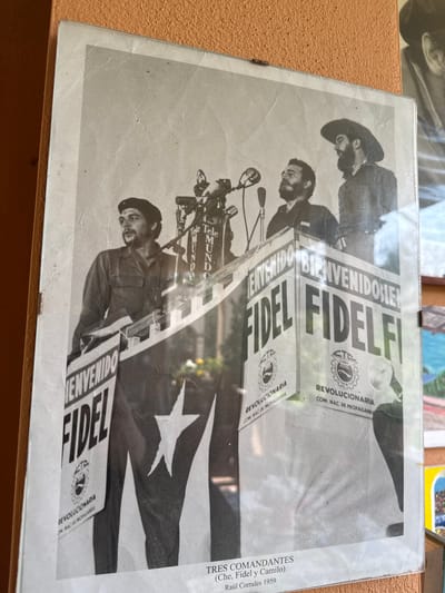 The image shows a black and white photograph, likely displayed in a frame. The photograph depicts three bearded men, identified in the text on the right as Che, Fidel, and Camilo, likely taken in 1959 by Raúl Corrales. The men are standing at a podium, holding signs that read "FIDEL BIENVENIDO" (Fidel Welcome). Cuban flags are also visible. The photograph is likely a historical document, perhaps on display in a museum or exhibition. The precise location within Rignano Flaminio, Italy cannot be determined based solely on the visual information in the image.