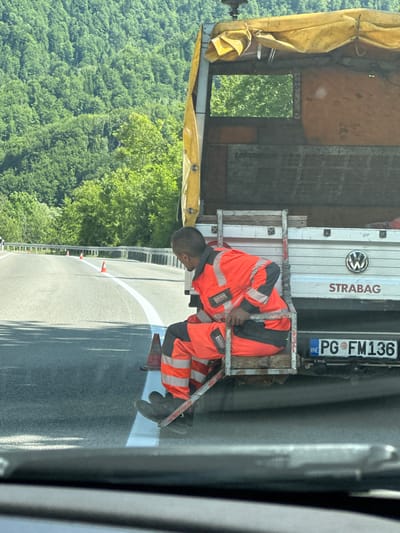 In the city of Crkvine, Montenegro, a man in a high-visibility orange and gray work suit is seated on the rear step of a white Volkswagen truck. The truck has a yellow tarp covering its bed, and the name "STRABAG" is visible on the side. The truck's license plate reads "PG FM136". The scene appears to be on a paved road with a white line being painted down the center. Several orange traffic cones are placed along the side of the road, suggesting ongoing road work. In the background, a dense forest covers a hillside, indicating a rural or mountainous setting. The perspective suggests the photo was taken from another vehicle traveling behind the truck.