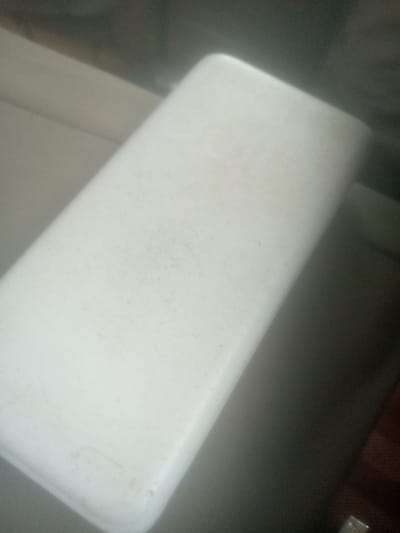 This media file shows a close-up, angled shot of a white, rectangular object with rounded edges. The object appears to be made of plastic and has some light scuff marks and faint smudges on its surface, suggesting it has been used. It is positioned on a textured surface, possibly a couch or upholstered furniture, which is a light beige color. A darker, possibly wooden surface is visible to the right of the white object, and a dark shadow obscures the background.
The lighting suggests an indoor environment, with soft light illuminating the top surface of the object, creating a gentle gradient of light and shadow across its form. There is no clear indication of the time of day or weather. The overall impression is of a simple, everyday object in a domestic setting. No people are visible, nor is there any text or discernible logos on the object. The image is slightly out of focus, with the most detail concentrated on the upper surface of the white object. The hints provided offer closer views of the surface texture and the transition between the white object and the darker background elements, reinforcing the interpretation of the scene.