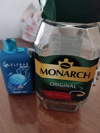 The image shows a close-up of a glass jar of Monarch Original Intense coffee and a blue Elf Bar disposable vape device. The coffee jar is partially filled with coffee grounds and has a green lid. The Elf Bar vape has a blue mouthpiece and is printed with "ELFBAR GH2800B" and an image of a blue orb with lines radiating from it, and "BLUEBERRY PEAR" written below. The objects are placed on a wooden surface, likely a table. In the blurred background, a white plastic bag with red circular patterns is visible. There is no indication of the city of Gorodovikovsk, Russia, or any people in the image. The scene appears to be an indoor setting.