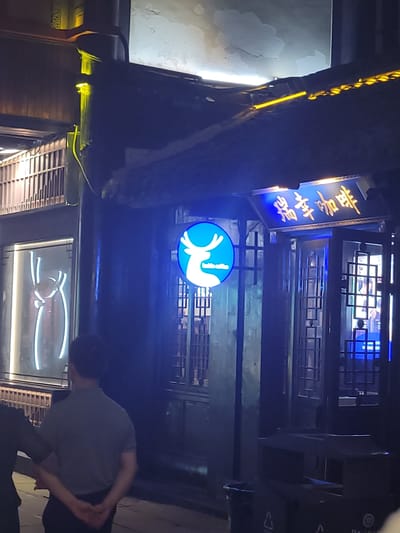 The image shows a night scene outside a coffee shop in Wuhou District, China. A man in a gray polo shirt and dark pants stands with his back to the camera, his hands clasped behind him. Another person, mostly obscured, is to his left. The coffee shop has a traditional Chinese architectural style with dark wooden beams and latticework. A bright blue neon sign with a white deer silhouette is displayed prominently. Above the entrance, a golden sign with Chinese characters is illuminated. The street is dimly lit, with yellow and blue lights from the shop and surrounding buildings. A black trash receptacle is visible in the foreground.
