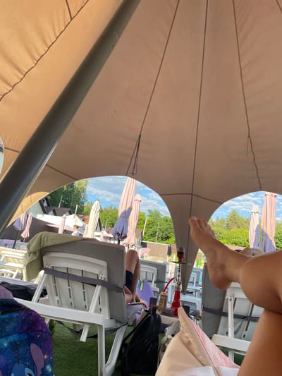 The image depicts a scene at a resort in Zazymia, Ukraine, under a large, beige, canopy-like structure supported by a thick, grey pole. The perspective is low, looking up from a reclined position. In the foreground, a person's bare feet are visible, with one leg extended towards the viewer. The feet are resting on a cushioned surface, possibly a beanbag or pillow, which is pink and white. To the left, a lounge chair with a grey cushion and a green towel draped over it is visible. Further back, other lounge chairs and closed umbrellas are arranged on what appears to be artificial grass. In the middle ground, a red hookah pipe with a glass and hose is placed on a small table or surface. Several beverage cans, including one with a yellow label that reads "JUICE," are also present. Beyond the seating area, there are buildings with dark roofs and several tall green trees. The sky is visible through the gaps in the canopy and is blue with scattered white clouds. In the background, a person in a white t-shirt and dark shorts is walking away. The overall impression is a relaxed, outdoor setting, likely a poolside or beach resort.