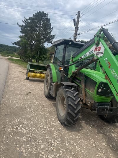 A green Deutz-Fahr tractor with an attached front loader and a rear-mounted John Deere agricultural implement is parked on a paved road. The tractor's tires are large and knobby, suggesting agricultural use. An electrical pole and overhead wires are visible in the background, along with a tree. The road surface is uneven and appears to be made of asphalt with embedded stones. The scene is outdoors under a cloudy sky. There is no visible evidence to pinpoint the location in Topchii, Bulgaria.