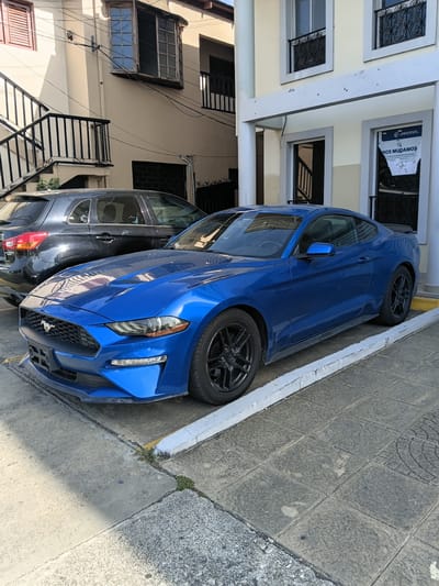 A blue Ford Mustang and a dark SUV are parked on a city street in Puerto Plata, Dominican Republic. The Mustang is in the foreground, angled towards the viewer, its front end slightly raised by a parking curb. The SUV is parked behind it, mostly obscured. The street is paved with concrete and interlocking tiles, and a building with a pale yellow facade and white trim rises behind the cars. A sign on the building's window reads "NOS MUDAMOS" which translates to "WE ARE MOVING" in Spanish. An exterior staircase with black railings is visible to the left of the building. The scene is bathed in sunlight, casting shadows on the street.