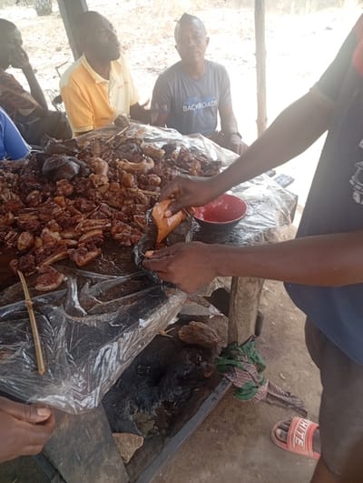 The image shows a table covered with a plastic sheet, piled high with a large quantity of meat. The meat is arranged in a haphazard manner, with some pieces being larger than others. A red plastic bowl is on the table, and a person in a blue shirt with a "BACKROADS" logo is sitting in the background. A person in a yellow shirt is sitting in the background. A person dressed in a blue shirt is preparing to eat from the table. The scene is likely in a public marketplace, restaurant, or street food stand in Assai Sabongida, Nigeria, based on the provided coordinates. The image is likely taken from the perspective of a person standing near the table. The scene is focused on the table and the meat, but other people and objects are in the background. The lighting is bright. The image is well-composed and in focus. It captures the bustling energy and activity of a crowded marketplace. The image provides a glimpse into the daily life of people in this part of Nigeria. The foreground is dominated by the table covered in meat, while the background shows a group of people sitting or standing and interacting with the meat. The scene is likely set in an outdoor area as there is foliage in the background. The person taking the photo is likely an observer who is standing close to the table.