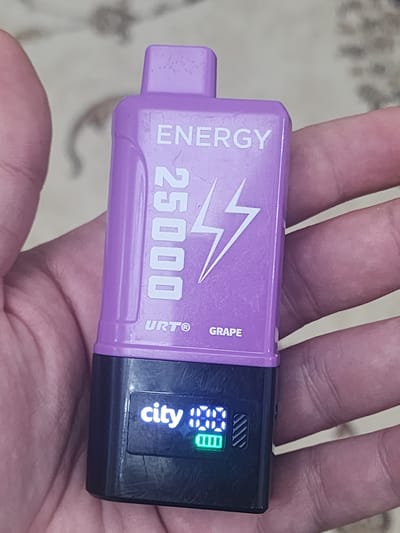The image shows a person's hand holding a purple and black electronic smoking device, commonly known as a vape. The device has text printed on it, including "ENERGY," "25000," "URT®," and "GRAPE." A small digital screen on the device displays the word "city" and a battery indicator with a green bar. The background appears to be a patterned surface. There is no visible indication of the city of Gudermes, Russia.