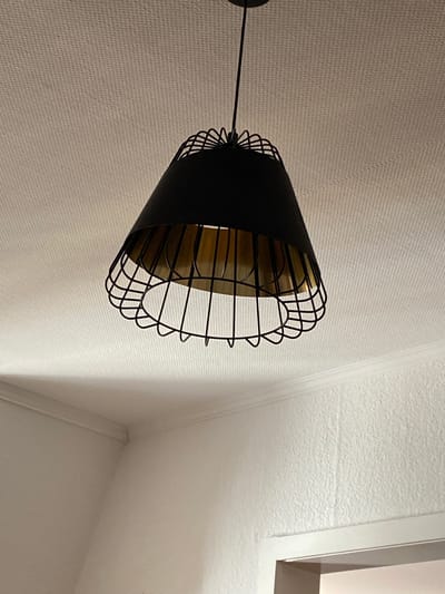A black pendant light with a conical shade and a wire cage is hanging from a textured white ceiling. The light fixture is in the center of the image, angled slightly to the right. The interior of the shade appears to be a golden color. The ceiling has a fine, linear texture. In the lower portion of the image, a white wall is visible, meeting the ceiling with a white molding. The wall also has a textured finish, appearing rougher than the ceiling. No people or animals are present in the image. The scene appears to be an indoor residential setting. This image was captured in Hildesheim, Germany.