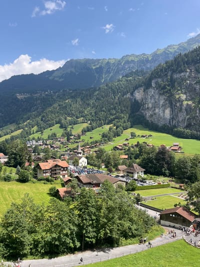 The image depicts a panoramic view of the mountain village of Wengen, Switzerland. In the foreground, a paved pathway runs horizontally, bordered by lush green grass and trees. Several individuals are walking along the pathway. Beyond the pathway, more trees and grassy slopes lead up to a cluster of buildings that form the village.
The village architecture features traditional Swiss chalets with wooden balconies and steeply pitched, terracotta-colored roofs. A prominent white church with a tall steeple stands at the center of the village, surrounded by a parking lot filled with cars. Numerous houses and smaller buildings are scattered throughout the village, nestled amongst the trees and on the terraced hillsides.
In the background, the landscape rises dramatically into steep, rocky mountains covered in dense green forests. The slopes are further accentuated by terraced fields and the sheer rock faces of cliffs. The sky above is a clear blue, dotted with a few white, fluffy clouds. The overall scene conveys a serene and picturesque mountain village setting.