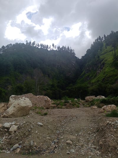The image depicts a mountainous landscape in Kalalengkio, Indonesia, under a cloudy sky. In the foreground, a dirt path is visible, littered with rocks and debris. Large boulders are scattered on either side of the path, with one particularly large, light-colored boulder dominating the left side of the frame. The middle ground features a dry, bare tree standing amidst greenery and more scattered rocks. The background is characterized by steep, densely forested mountainsides. The vegetation consists of numerous trees, with some patches of lighter green grass on the slopes. The overall scene suggests a natural, somewhat rugged environment, possibly with recent geological activity or land disturbance due to the presence of loose rocks and the unpaved path. No people are visible in the image.