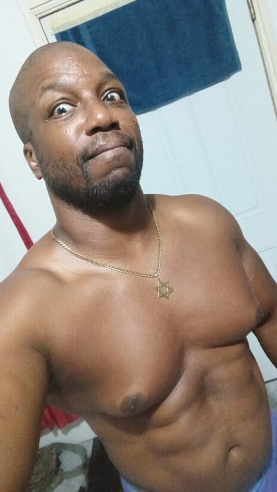 A bare-chested man with a bald head and a goatee looks upward with wide, surprised eyes. He is wearing a gold chain with a Star of David pendant. The background is a plain wall with a blue towel hanging on it. The man appears to be in a bathroom or bedroom. The scene is lit by overhead lighting, and there is a slight sheen of sweat on the man's forehead. The image is a selfie, taken from a low angle.