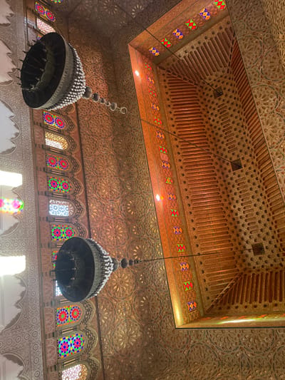 The image captures an upward view of an elaborately decorated interior within the city of Moulay Idriss Zerhoun, Morocco, likely the Mausoleum of Moulay Idriss I. The scene is dominated by a richly carved wooden ceiling and upper wall sections. The ceiling features intricate geometric and curvilinear patterns in warm brown and golden tones. Two large, dark-toned chandeliers, each adorned with numerous white, spherical elements, hang from chains. Along the upper left, a series of arched windows with colorful stained glass panels displaying geometric and floral motifs allow natural light to filter through, casting faint colored reflections. A horizontal decorative wooden frieze, containing smaller colored glass inserts, runs across the upper wall, transitioning into a recessed, coffered ceiling section also crafted from carved wood. No individuals are present in the static scene.
