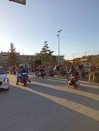 In the city of Arbia, Italy, an outdoor gathering of motorcyclists is captured in a scene bathed in the warm light of a late afternoon or early evening sun. The foreground is dominated by a white car parked on the left, its headlights illuminated. To its right, a blue motorcycle, with its rider wearing a white helmet and red shoes, is the central focus. The rider's legs are visible as they are positioned to ride.

A crowd of people and numerous motorcycles are visible in the midground and background, suggesting a rally or event. The motorcycles are diverse in make and model, with many appearing to be cruiser-style bikes. Riders and spectators, mostly men, are seen standing, sitting on their bikes, or walking. Many are wearing motorcycle gear, including helmets and leather vests, some adorned with patches.

The environment includes a paved ground surface, likely a parking lot or square, with long shadows cast by the sun. In the background, buildings with multiple stories, possibly apartments or offices, are visible. Trees, including pine trees, and a tennis court with a net are also present, indicating an urban or semi-urban setting. A street light with two fixtures stands tall against the clear blue sky, adding to the ambient lighting. The overall impression is a community event centered around motorcycles on a pleasant day.