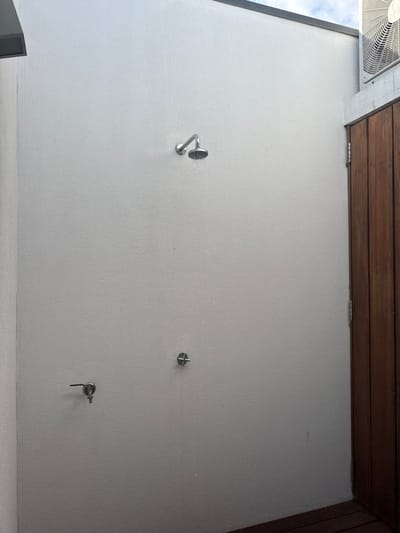 The scene depicts an outdoor shower area located in Grândola, Portugal. A textured light grey wall forms the primary background, extending from the bottom to the top of the frame. Approximately one-third down from the top and slightly left of the vertical center, a chrome-finished showerhead is mounted on this wall, actively dripping or flowing water. Below the showerhead, two chrome water control fixtures are installed: a lever-style faucet handle on the lower left, and a circular knob to its right.
To the right side of the image, a vertical section of dark brown wooden planks, consistent with a door or a partition wall, is visible, extending from the bottom upwards. In the upper right corner, an external air conditioning unit's white gridded fan cover is partially visible, mounted above the wooden structure and set against a light blue sky with scattered white clouds. The floor in the lower right corner appears to be dark wooden decking. No individuals are present in the image.