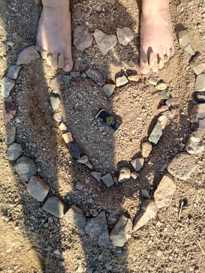This media file captures a close-up, overhead view of a heart shape formed by small rocks and pebbles on a sandy surface. Two bare feet, with small pebbles placed on the toes, are visible on the left side of the frame, suggesting a person is interacting with the arrangement. The feet are lightly dusted with sand.
In the center of the rock heart lies a small, rectangular object that appears to be a lighter. It is black with a gold design, possibly a playing card symbol like an ace of spades or a stylized crest.
The scene is set outdoors, indicated by the natural light and the presence of sand and rocks. The lighting suggests it is daytime, with distinct shadows cast by the rocks and the feet, implying a sunny environment. The overall tone is relaxed and possibly playful, given the arrangement of rocks and the casual placement of the feet.
There is no visible text in the media file itself, other than the potential design on the lighter, which is not clearly legible. The location context provided is Yuzhny, Kazakhstan, which aligns with the earthy, outdoor setting. The texture of the sand and the variety of rock shapes and colors are notable details.