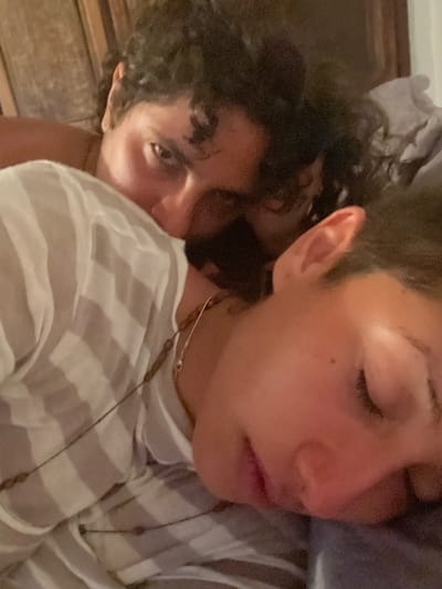 Two people are lying down. The person on the right has short hair, closed eyes, and is wearing a white and gray striped shirt with two necklaces. The person on the left has curly dark hair and is looking at the camera. They are close together, possibly cuddling. The lighting is dim.